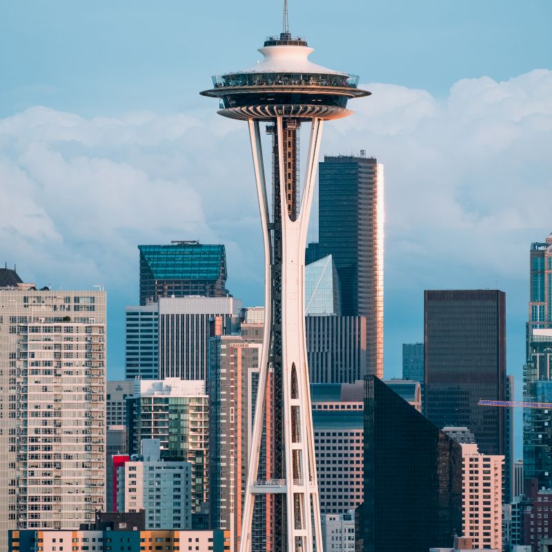 The image shows a cityscape with a tall, iconic structure in the foreground, surrounded by modern buildings and skyscrapers, under a clear sky.