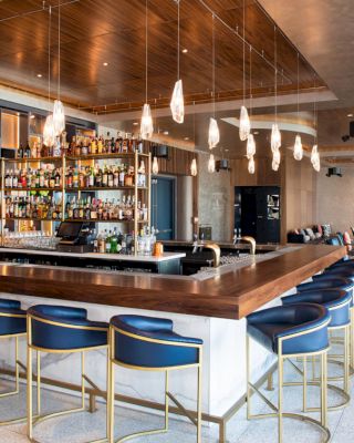 The image shows a modern bar with blue and gold bar stools, a well-stocked liquor shelf, pendant lights, and a seating area in the background.
