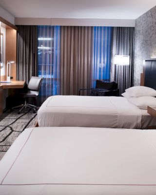 This image shows a modern hotel room with two twin beds, a desk area, a flat-screen TV, and large windows with curtains.