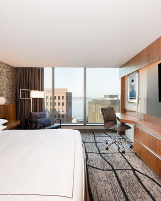 A modern hotel room with a large bed, TV, desk, chair, armchair by the window, and a city view through floor-to-ceiling windows.