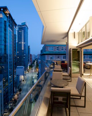 A modern balcony overlooking a cityscape at dusk, with high-rise buildings, outdoor seating, and a well-lit interior space.