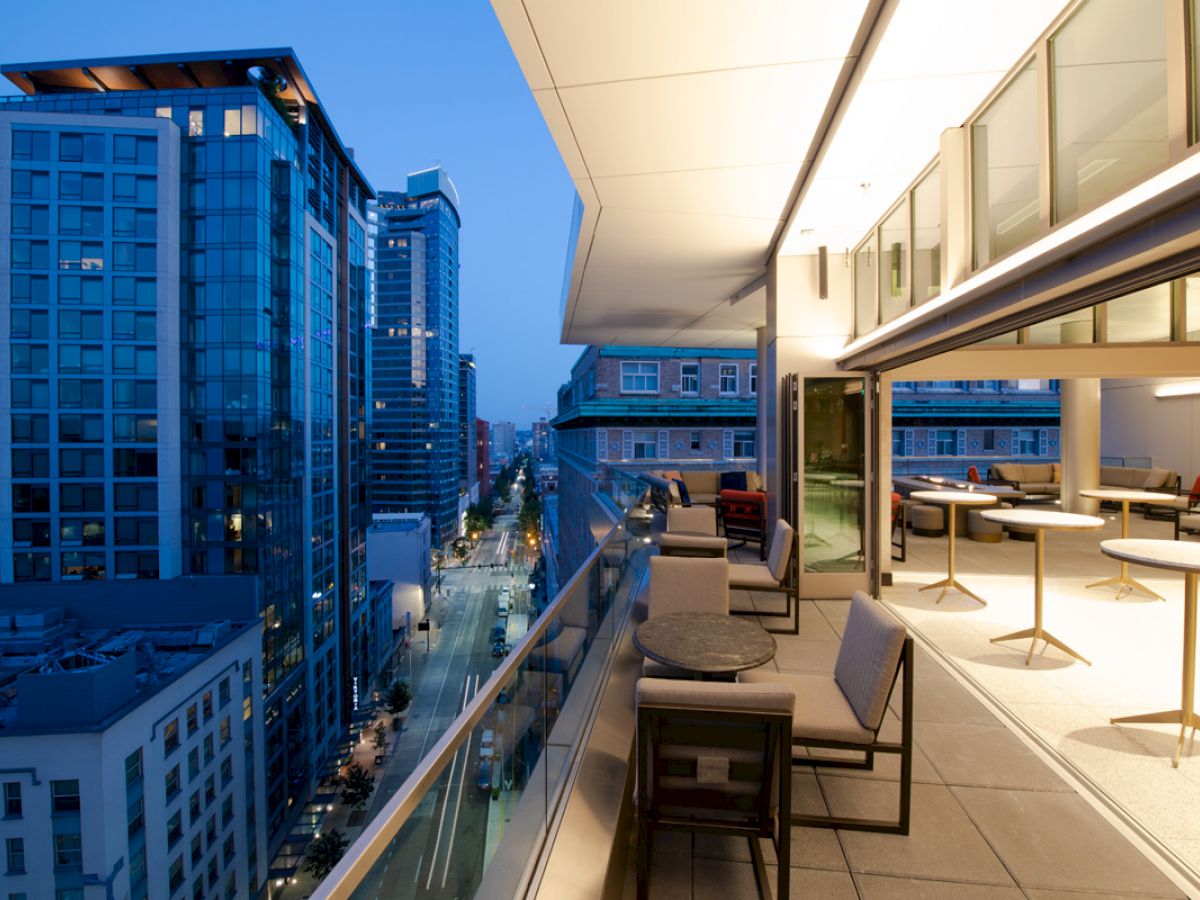 A modern cityscape view from a balcony at dusk, showcasing tall buildings and a stylish outdoor seating area with tables and chairs.