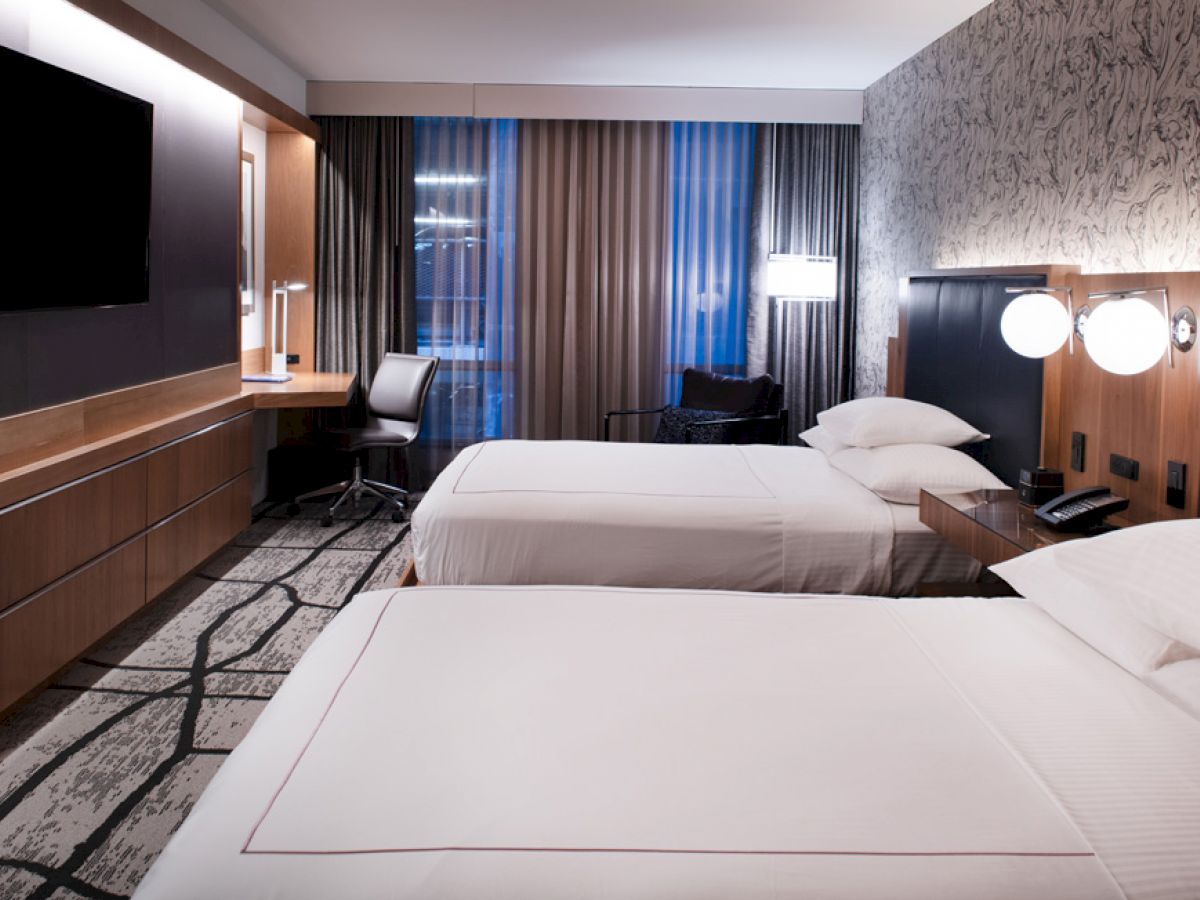 A modern hotel room with two neatly made beds, wall-mounted TV, work desk, and ambient lighting; the decor is sophisticated with patterned wallpaper.