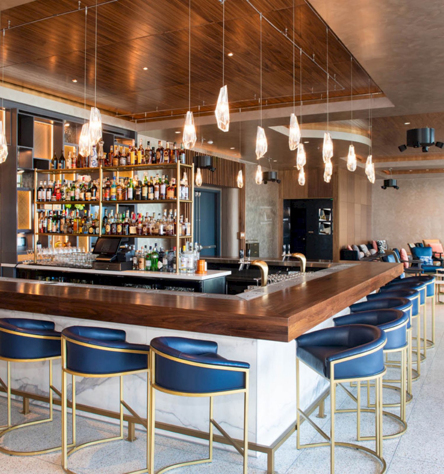 This image shows a modern bar with high seating chairs, a fully stocked bar, hanging lights, and a seating area in the background.