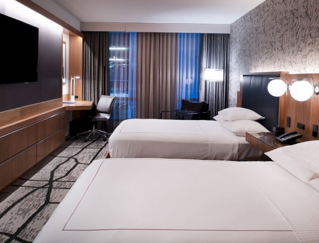This is a modern hotel room with two beds, a large TV, a desk with a chair, and contemporary decor. The room has large windows with curtains.
