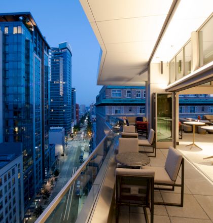 The image shows a modern cityscape with tall buildings, viewed from a balcony with tables, chairs, and an open-air dining area.