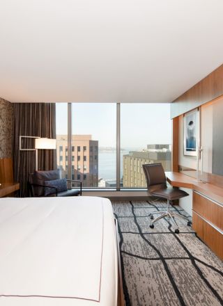 A modern hotel room with a large bed, flat-screen TV, desk, chair, and a window offering a city view.