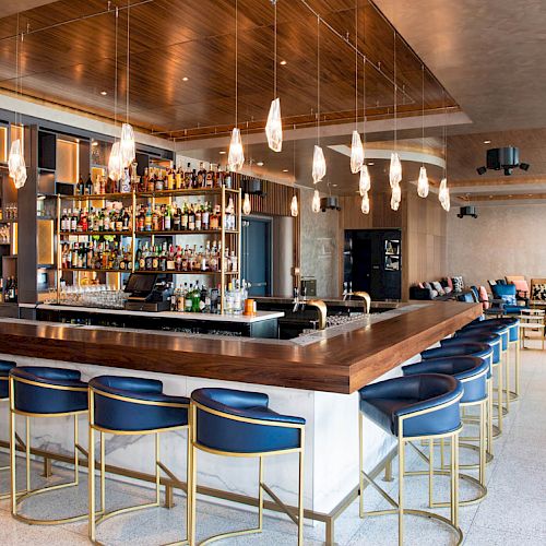 A modern bar with a row of blue stools, a well-stocked liquor shelf, hanging lights, and a lounge area with seating in the background.