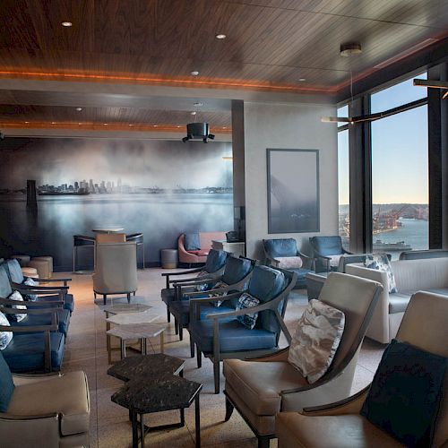 The image shows a modern lounge with large windows, numerous chairs, tables, and a mural on the wall, overlooking a scenic waterfront city view.