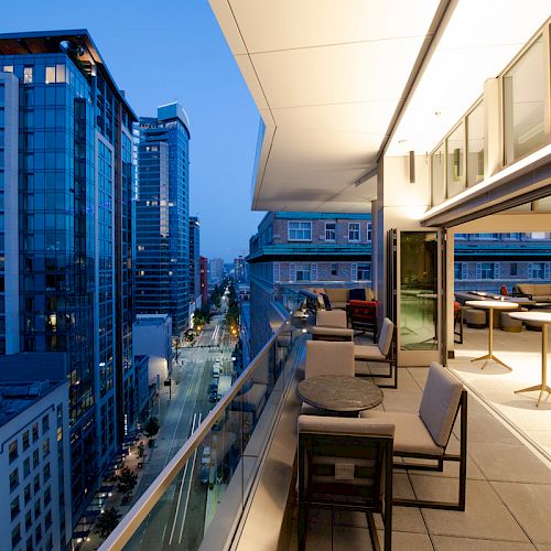 This image shows a modern cityscape view from a balcony with outdoor seating at dusk. Tall buildings line the street below, illuminated by streetlights.