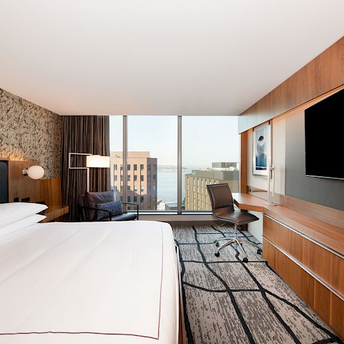 The image shows a modern hotel room with a large bed, a desk with a chair, and a wide-screen TV. There's a view of buildings outside the window.