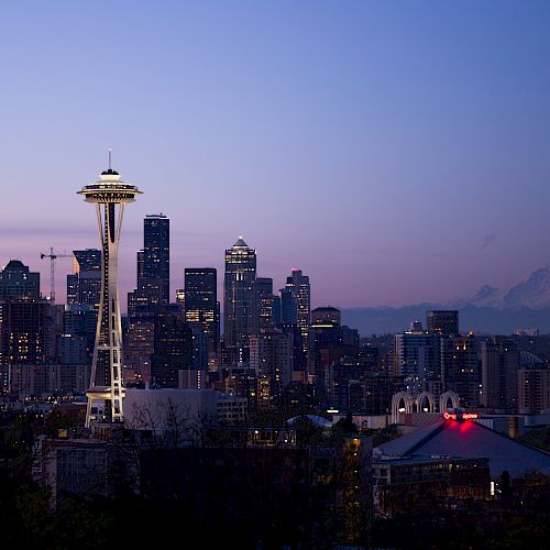 A nighttime cityscape featuring a prominent needle-like tower with a mountainous backdrop and several illuminated buildings against a twilight sky.