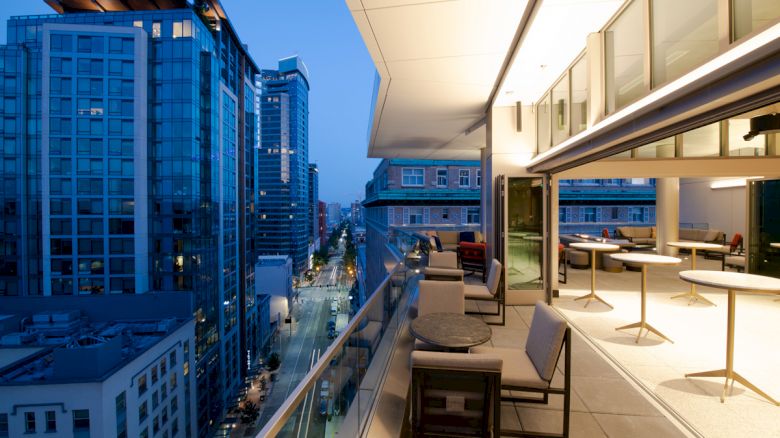 The image depicts a modern cityscape at dusk from a high balcony of a building with seating and tables, overlooking a street with tall buildings.