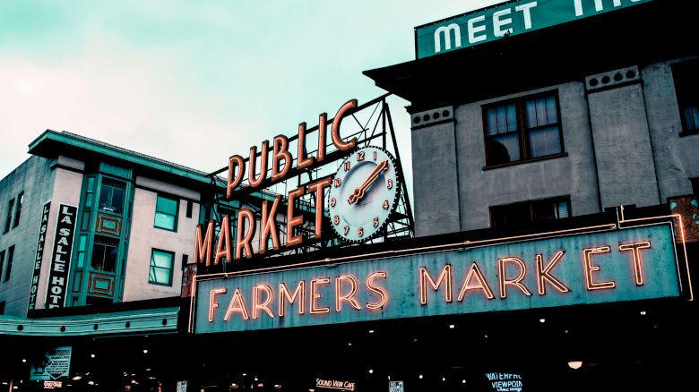 The image shows the iconic neon signs of the Pike Place Market in Seattle, reading "Public Market" and "Farmers Market" with a classic clock.
