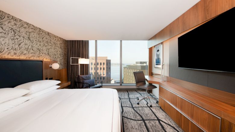 A modern hotel room with a large bed, a desk with a chair, wall-mounted TV, and a view of buildings and water through the window.