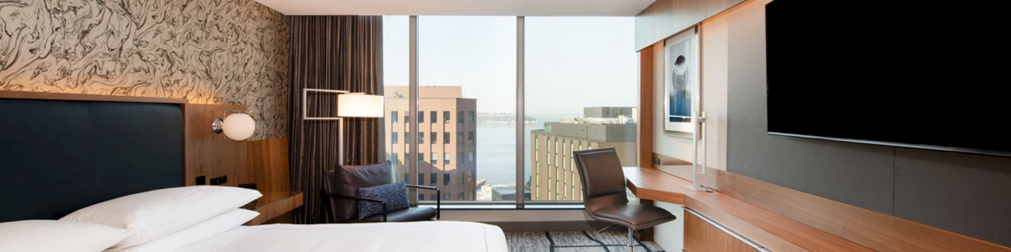A modern hotel room with a large bed, wall-mounted TV, desk, chair, and a window with a city view and water in the distance.