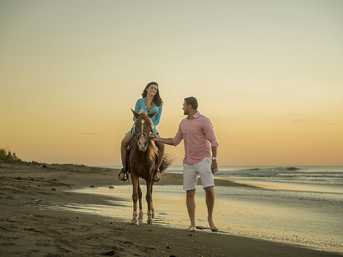 A woman is riding a horse on the beach while a man walks beside her, holding the horse's reins, during a sunset.