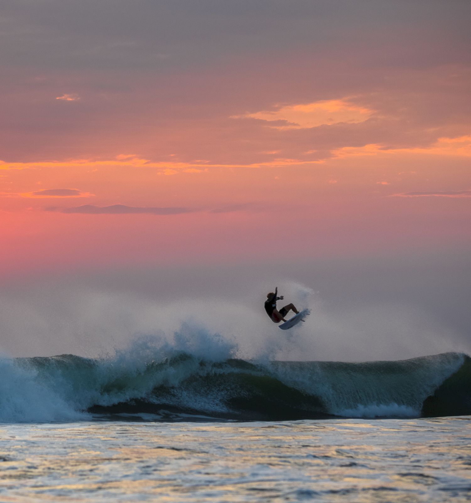 A surfer is riding a wave during a beautiful sunset, with the sun low on the horizon and the sky filled with orange and purple hues.